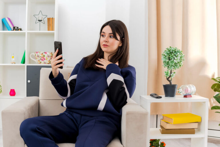 impresionada-joven-bonita-mujer-caucasica-sentada-sillon-sala-estar-disenada-sosteniendo-mirando-telefono-movil-manteniendo-mano-pecho (1)